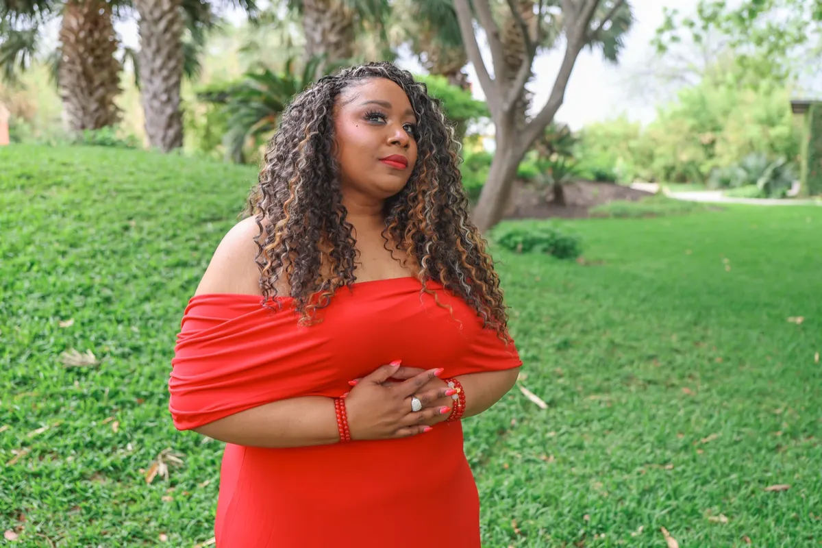 Andrea “Vocab” Sanderson in a garden, hands clasped, gazing upward in a red off-the-shoulder dress